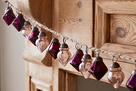 The Ekiti Bauble Garland - Ruby & Copper by nkuku features geometric, recycled glass ornaments with metal caps, strung on twine for a festive, vintage look—perfect for adding cozy charm to your mantelpiece.