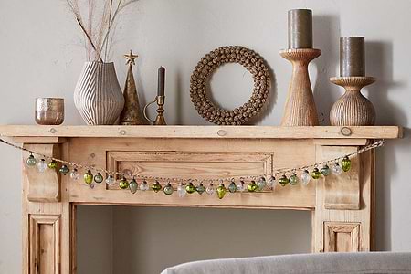 A light wooden mantelpiece decorated with vases, candles, a round beaded mirror, and the nkuku Ekiti Bauble Garland in mixed green and clear. Textured ceramics, recycled glass, and dried grasses add cozy festive charm.