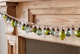 The Ekiti Bauble Garland - Mixed Green & Clear by nkuku decorates a rustic, natural-wood mantel with vintage-style recycled glass ornaments in green and clear, each hung with twine to highlight the wood’s visible grain and knots.