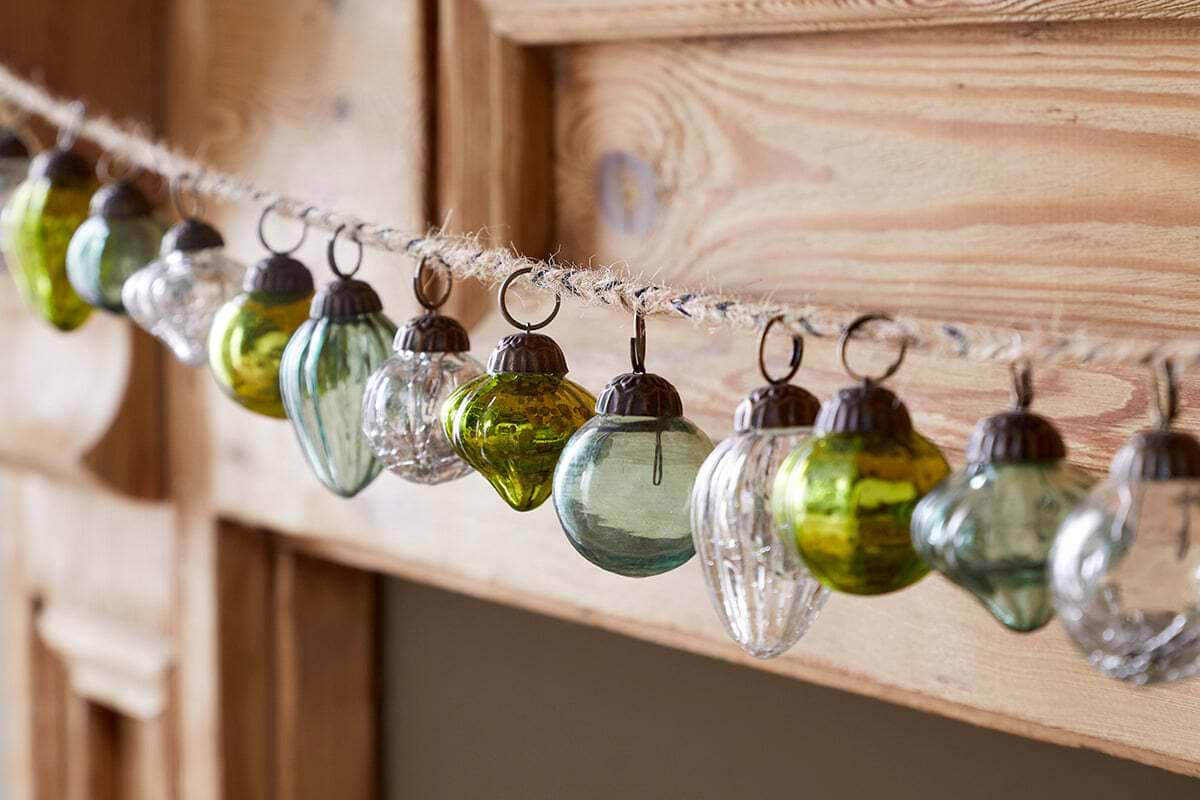 The Ekiti Bauble Garland - Mixed Green & Clear by nkuku features small recycled glass ornaments in green, clear, and silver, with round and ribbed shapes, all hanging from twine—ideal for rustic festive décor against a light wood background.