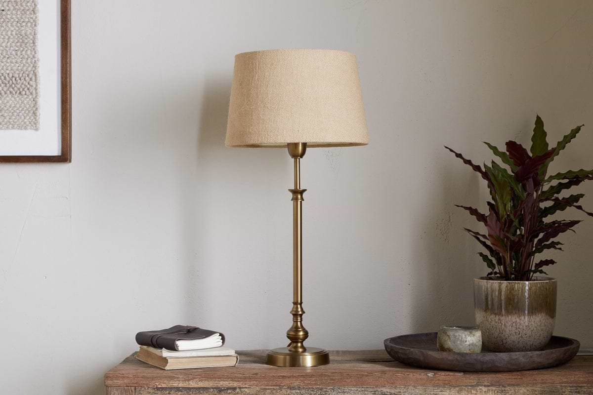 The nkuku Ehadi Antique Table Lamp in antique brass sits elegantly on the table, bringing a stylish and classic touch to your space.