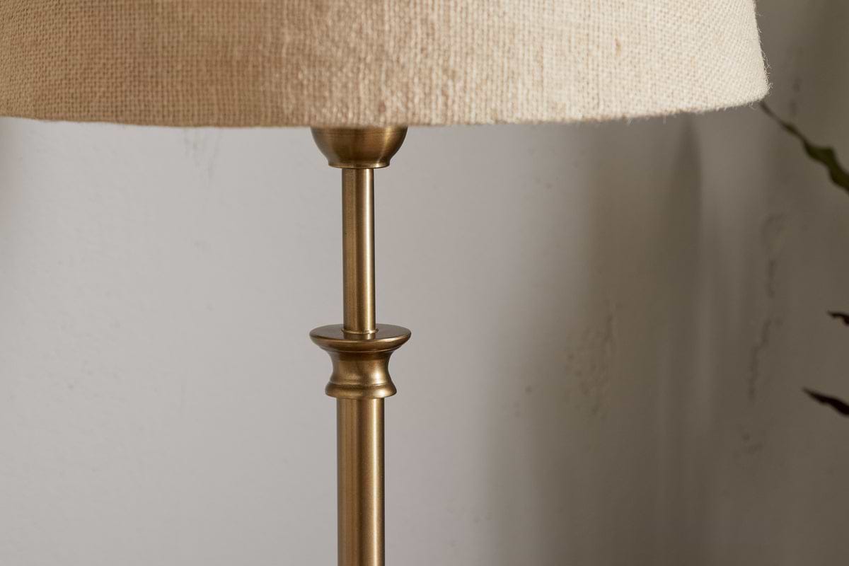 A close-up shows the upper part of the nkuku Ehadi Antique Table Lamp in antique brass, featuring a beige textured fabric shade against a light-colored wall.