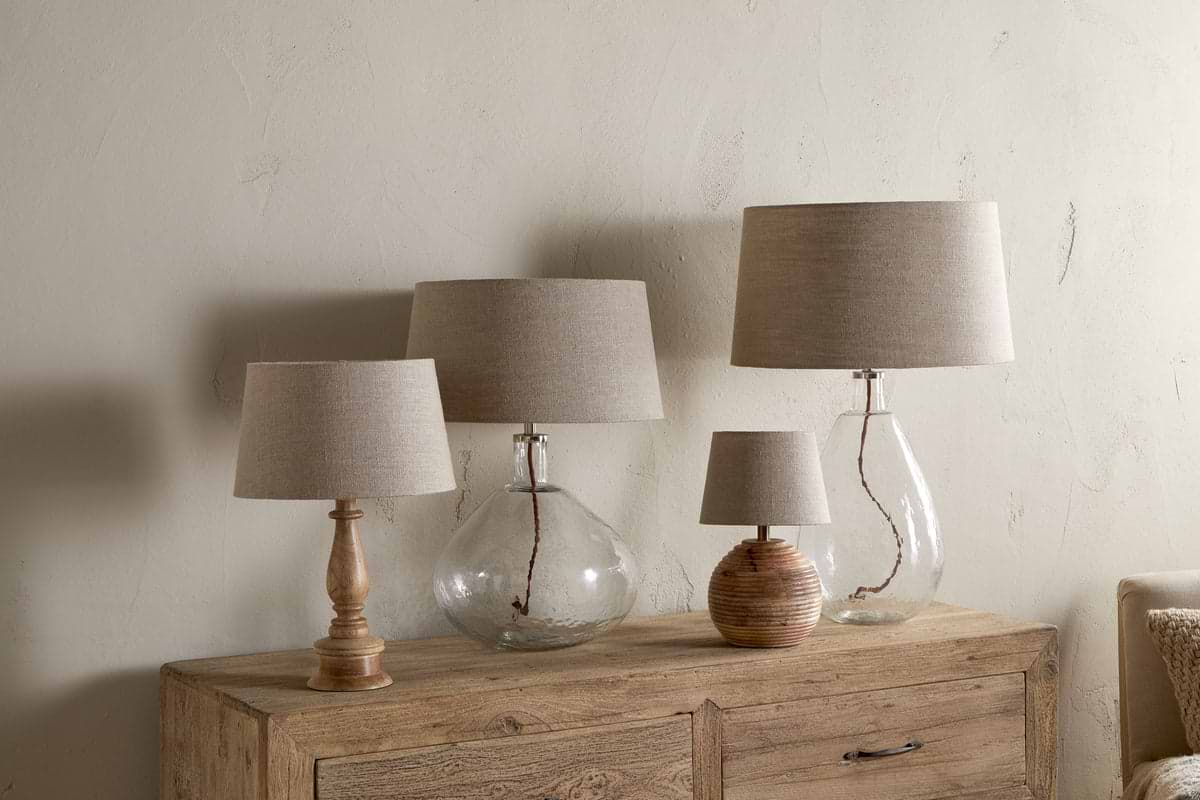 Four Embaata Linen Lampshades - Natural by nkuku, with textured linen, sit on a wooden sideboard—two have clear glass bases, one a wooden base, and another a ribbed round base. The neutral wall highlights their artisanal look.