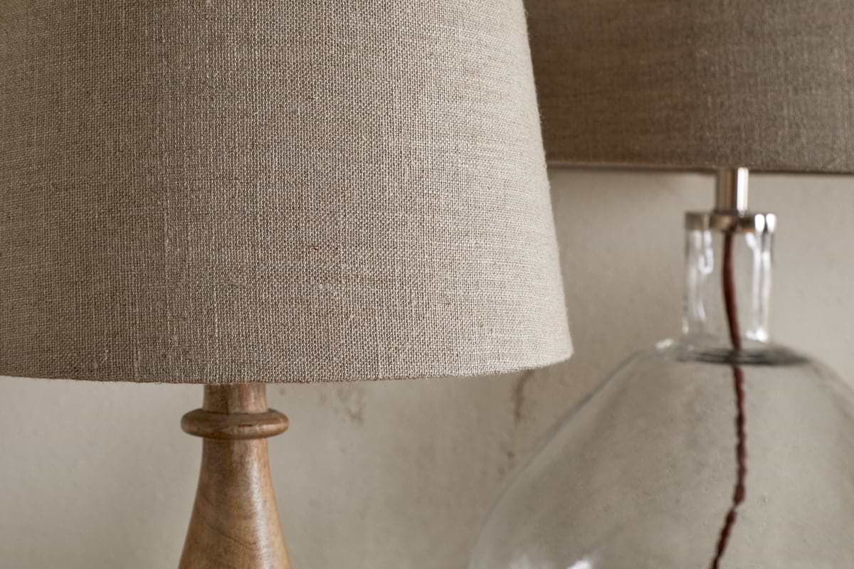 Close-up of two nkuku Embaata Linen Lampshade - Natural lamps: one with a wooden base, the other with a clear glass base—both perfect as bedroom lights against a neutral wall.