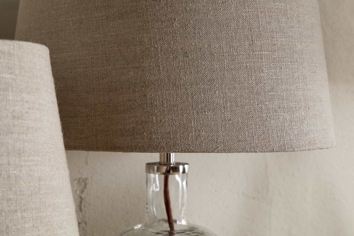 A close-up of the nkuku Embaata Linen Lampshade - Natural, featuring a textured linen shade and transparent glass base, sits beside a similar bedroom lamp against a neutral wall.