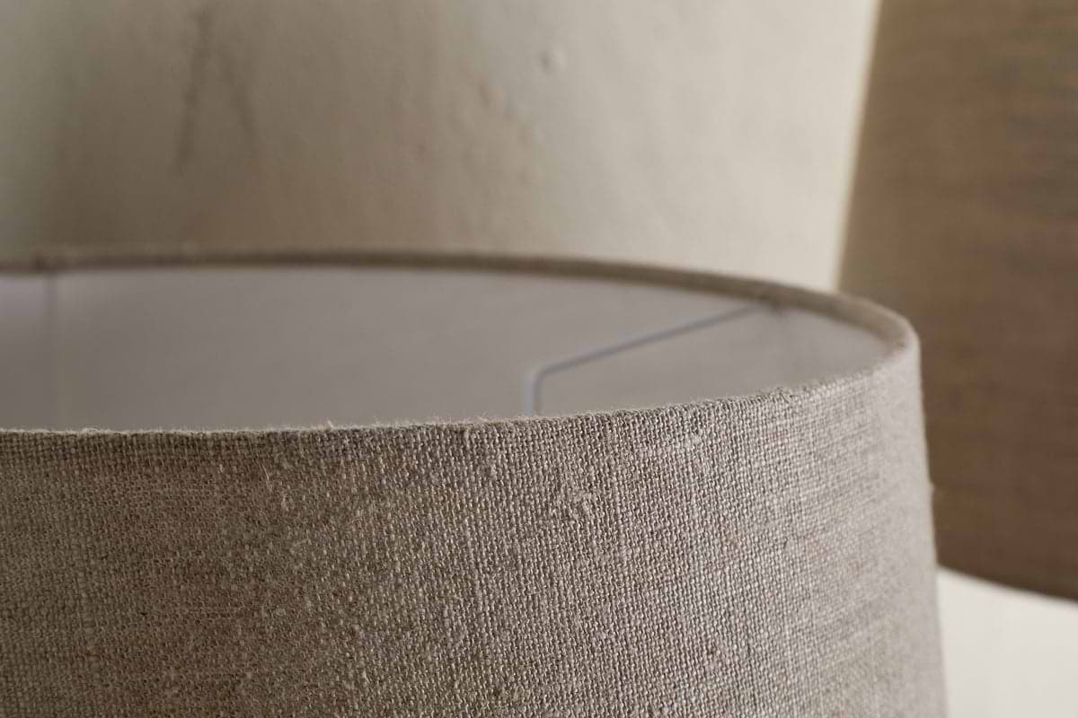Close-up of the nkuku Embaata Linen Lampshade in Natural, highlighting its beige textured linen and inner metal frame. A second lampshade is blurred in the background, ideal for soft bedroom lighting against a neutral wall.