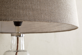 Close-up of the nkuku Embaata Linen Lampshade - Natural, featuring a textured linen shade and clear glass base, ideal for bedroom lighting, set against a light, neutral background.