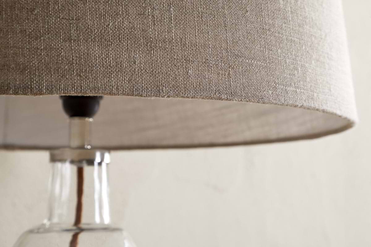 Close-up of the nkuku Embaata Linen Lampshade - Natural, featuring a textured linen shade and clear glass base, ideal for bedroom lighting, set against a light, neutral background.