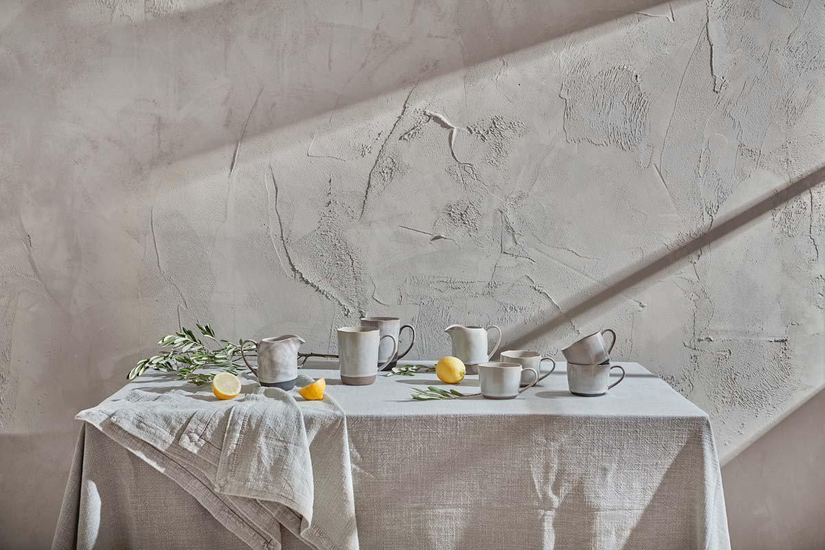 A table set with the nkuku Edo Mug - Large - Slate, ceramic pitchers, lemons, and olive branches sits in soft sunlight. Shadows play across a pale textured wall, creating a minimalist yet rustic atmosphere.