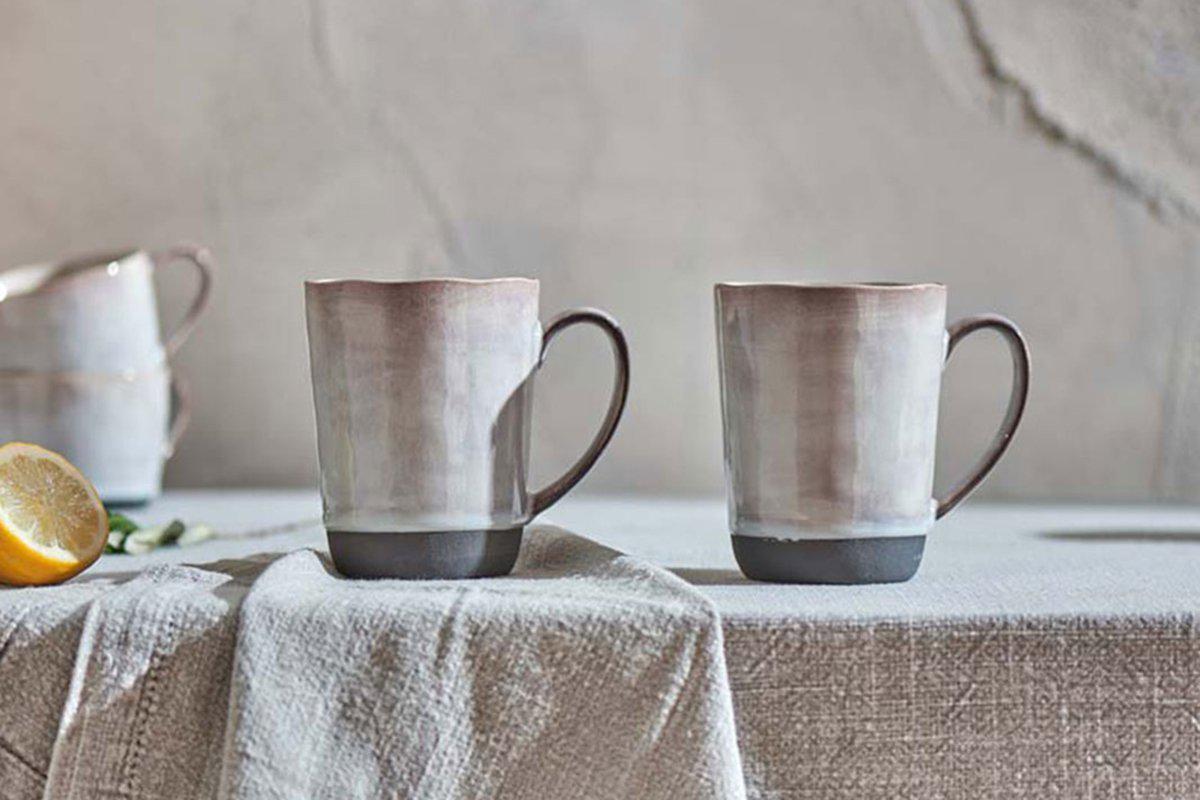 Two nkuku Edo Mug - Large - Slate mugs with a dark glaze and off-white finish rest on a linen tablecloth beside a lemon slice.