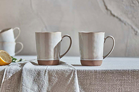 The nkuku Edo Mug - Large - Terracotta (Set of 2) features scalloped rims and a soft ceramic glaze, shown stacked with a lemon half on a textured cloth against a rustic, terracotta-hued cracked wall in a softly lit, minimalist scene.