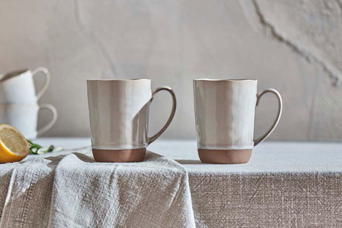 The nkuku Edo Mug - Large - Terracotta (Set of 2) features scalloped rims and a soft ceramic glaze, shown stacked with a lemon half on a textured cloth against a rustic, terracotta-hued cracked wall in a softly lit, minimalist scene.