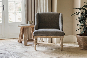 The nkuku Elbu Deconstructed Jacquard Weave Armchair in Charcoal with wooden legs sits on a stone tile floor next to a rustic mango wood side table holding a mug and book, while sunlight streams through glass-paneled double doors.