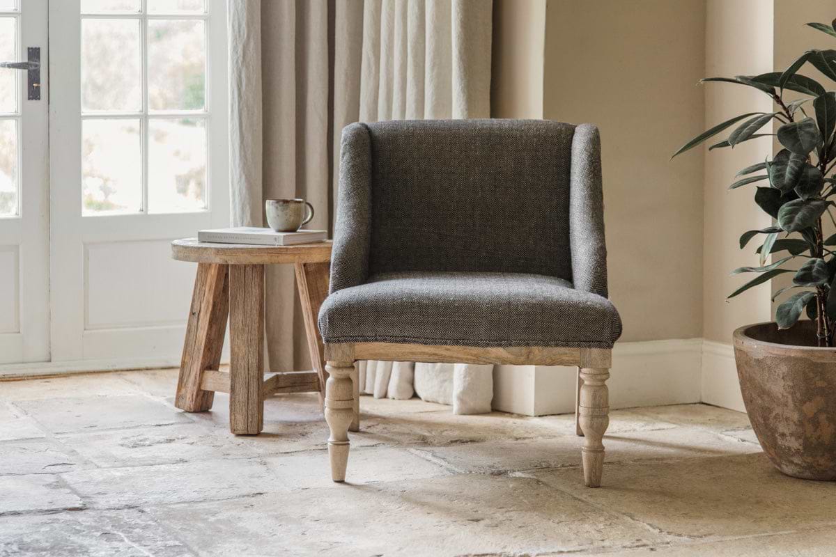 The nkuku Elbu Deconstructed Jacquard Weave Armchair in Charcoal with wooden legs sits on a stone tile floor next to a rustic mango wood side table holding a mug and book, while sunlight streams through glass-paneled double doors.