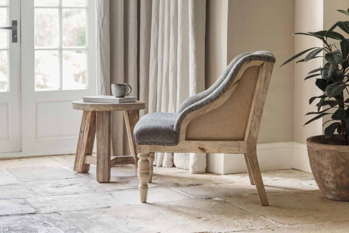 The nkuku Elbu Deconstructed Jacquard Weave Armchair in charcoal sits on a stone floor beside a round side table with a mug and book. Beige curtains frame French doors, and a potted plant rests in the corner next to the chair.