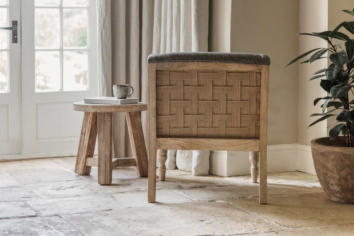 A cozy nook with nkuku’s Elbu Deconstructed Jacquard Weave Armchair in charcoal, a round wooden side table with a book and mug, stone tile floor, leafy potted plant, and sunlight streaming through white-paned French doors and curtains.