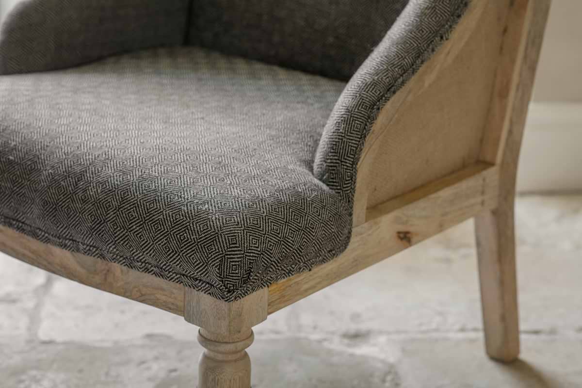 A close-up of the nkuku Elbu Deconstructed Jacquard Weave Armchair - Charcoal, crafted from light mango wood with hand-upholstered, textured charcoal gray jacquard fabric, shown on a pale stone or tile floor.