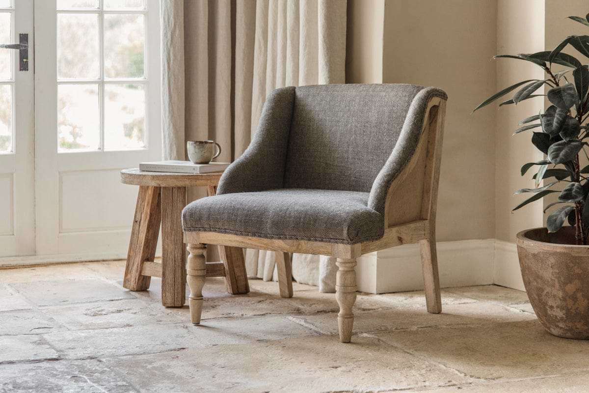 A cozy nkuku Elbu Deconstructed Jacquard Weave Armchair in Charcoal with wooden legs rests on stone tiles beside a rustic mango wood side table holding a mug and book. Sunlight pours through French doors, highlighting the neutral-toned space and potted plant.