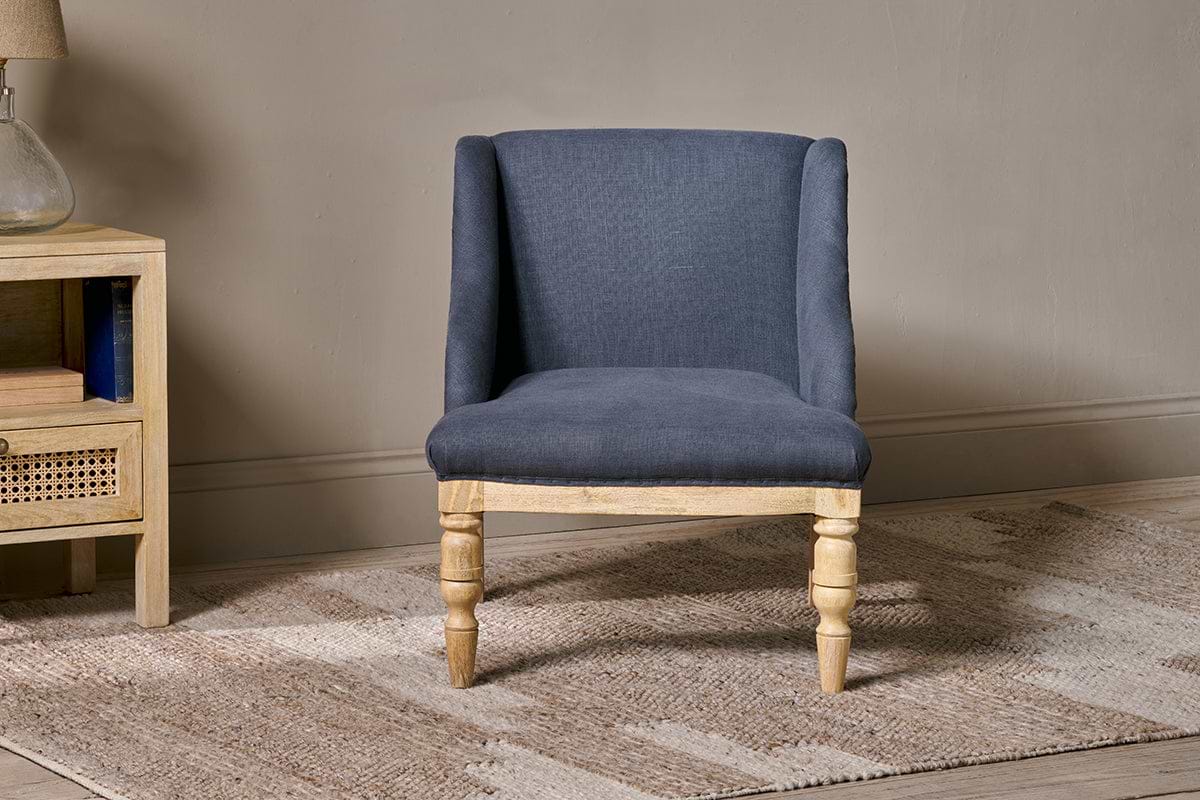 The nkuku Elbu Deconstructed Linen Armchair in charcoal, with lightly padded legs and a deep linen seat and backrest, sits on a beige woven rug. To the left, a light wood nightstand holds books below and a lamp on top.