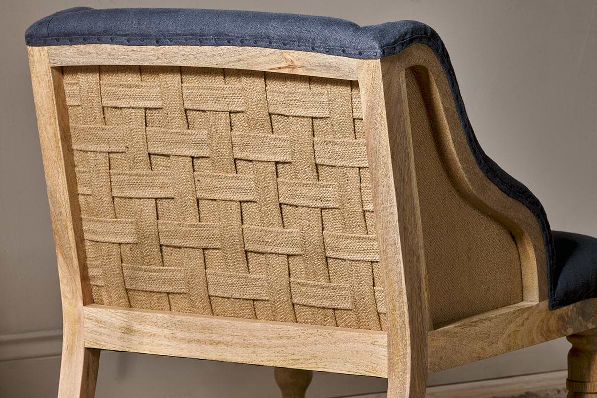 A close-up of the Elbu Deconstructed Linen Armchair by nkuku highlights its light mango wood, exposed woven burlap back, and charcoal linen fabric along the edges and seat—showcasing rustic charm with a modern twist.