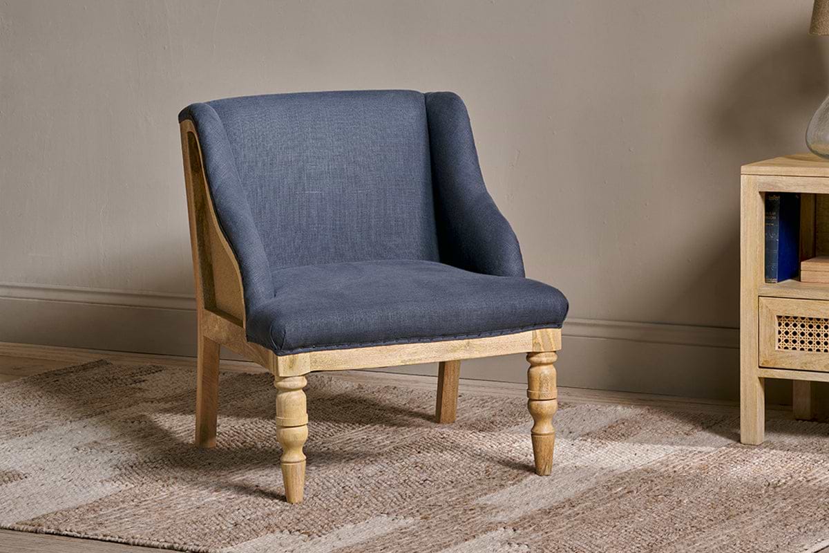 The nkuku Elbu Deconstructed Linen Armchair - Charcoal, featuring a dark linen upholstery and mango wood frame, sits atop a textured beige rug with a wooden side table nearby holding books and a woven basket against a neutral wall.