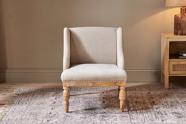 The nkuku Elbu Deconstructed Linen Armchair in Stone features a curved back and nailhead trim, wooden legs, and sits on a light patterned rug. Behind it: a tan wall, natural mango wood side table, and brown ceramic base lamp.