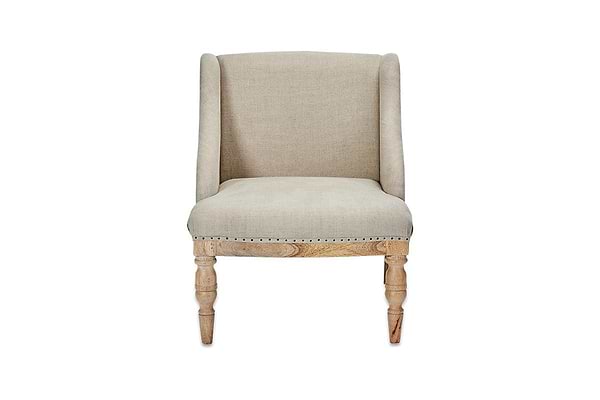 The nkuku Elbu Deconstructed Linen Armchair - Stone features beige linen upholstery, a curved backrest with winged sides, light mango wood turned legs, and nailhead trim along the bottom edge. It is shown against a plain white background.