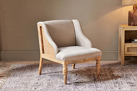 The Elbu Deconstructed Linen Armchair - Stone by nkuku, with light beige linen upholstery and a natural mango wood frame featuring turned front legs, sits on a patterned rug in a cozy room with neutral walls and wooden furniture.