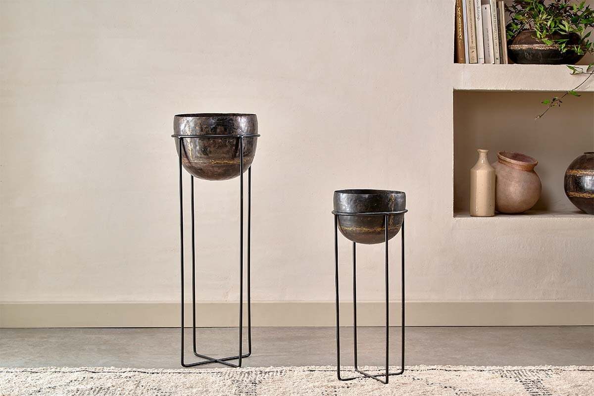 Two Endo Reclaimed Iron Planter Stands by nkuku, featuring shiny black bowls and slender legs, sit on a light rug against a beige wall. A built-in shelf with vases and jars completes the minimal, modern space.