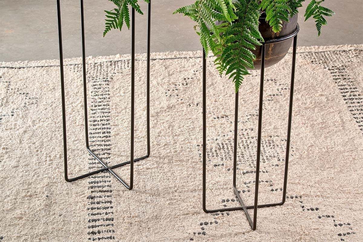 Two nkuku Endo Reclaimed Iron Planter Stands with geometric black legs display green ferns on a beige, natural woven rug with small black dots. The textured rug contrasts the gray concrete floor beneath.