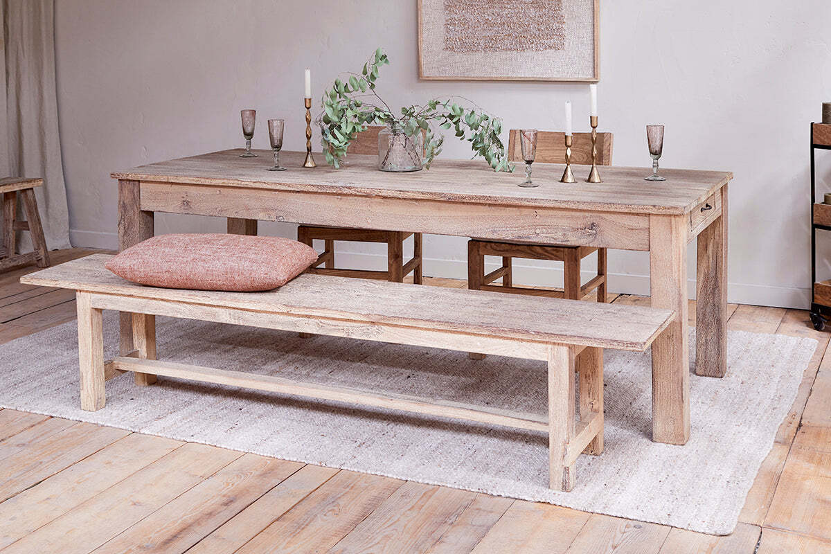 The nkuku Fioni Reclaimed Wood Bench – Natural sits on a light rug beside a rustic dining table. The bench features a brown cushion, while candlesticks, goblets, and a vase with green foliage decorate the table amid cozy neutral décor.