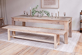The Fioni Reclaimed Wood Bench by nkuku pairs with a rustic dining table atop a light woven rug. Candlesticks, goblets, and a glass vase with greenery complete the timeless, neutral farmhouse look.