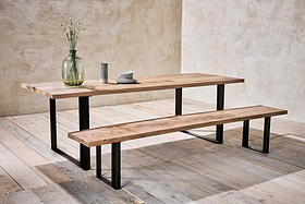 A dining set featuring a long wooden table and the nkuku Fia Bench with black angular legs sits on pale wood flooring. On the table are a glass vase with dried flowers, a cup, bowl, and plates against textured beige walls.