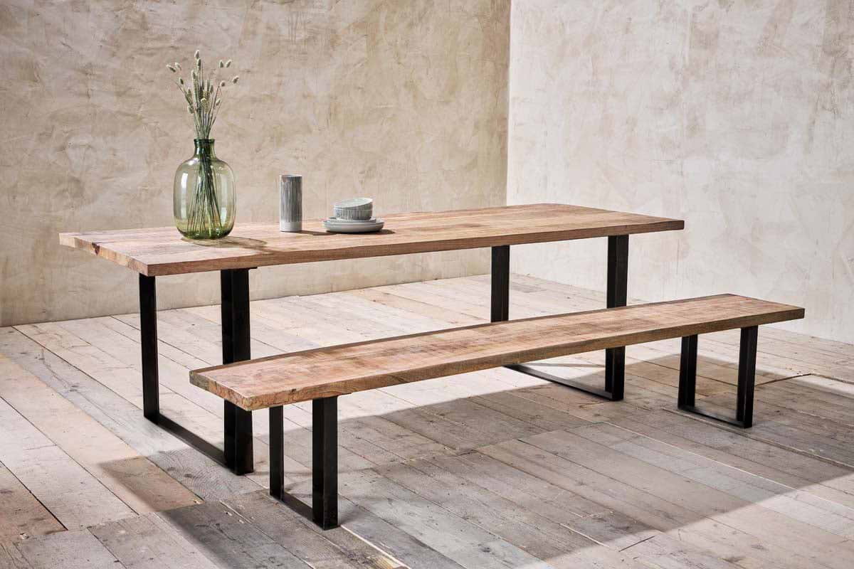 A dining set featuring a long wooden table and the nkuku Fia Bench with black angular legs sits on pale wood flooring. On the table are a glass vase with dried flowers, a cup, bowl, and plates against textured beige walls.