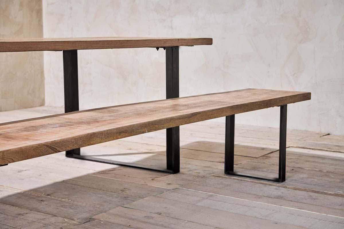 A close-up of the minimalist Fia Bench by nkuku, featuring a thick mango wood top and bold angular legs, set on rustic wooden floorboards against a textured light beige wall.