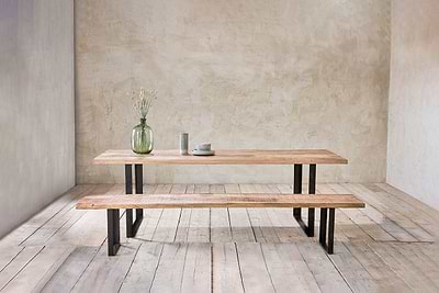 The nkuku Fia Bench, crafted from mango wood with angular legs, pairs with a matching table. On top: a green glass vase with dried flowers and ceramic dinnerware. The minimalist room features textured walls and wooden floors.