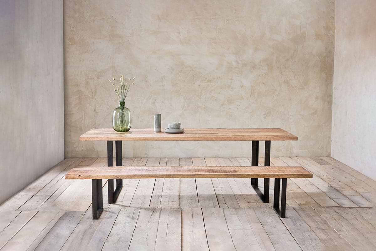 The nkuku Fia Bench, crafted from mango wood with angular legs, pairs with a matching table. On top: a green glass vase with dried flowers and ceramic dinnerware. The minimalist room features textured walls and wooden floors.