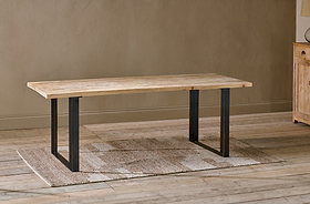 The nkuku Fia Dining Table, crafted from sustainable mango wood with a light natural finish and black iron base, stands on a textured beige rug over wooden floors against a light brown wall, with part of a wooden cabinet visible on the right.