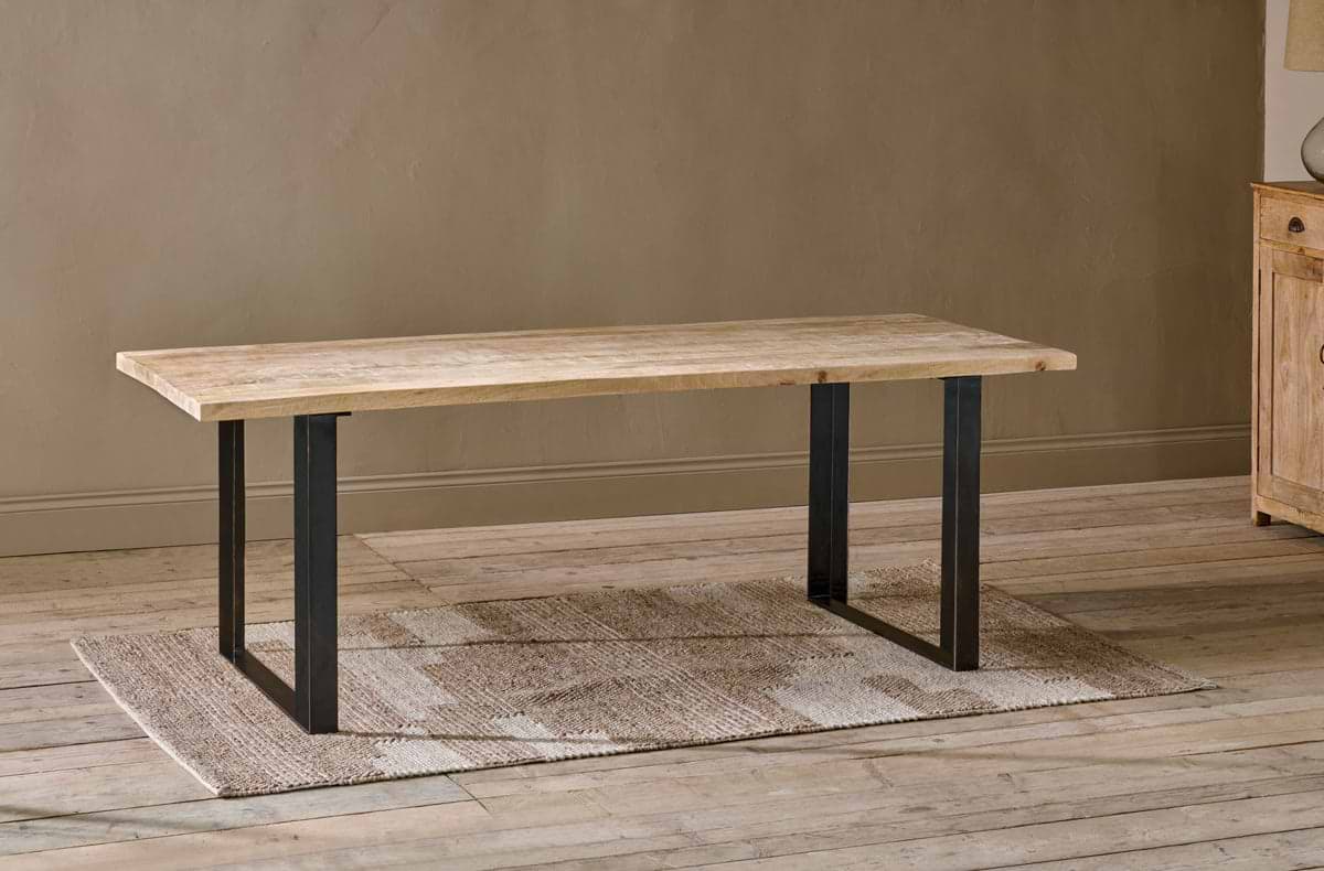 The nkuku Fia Dining Table, crafted from sustainable mango wood with a light natural finish and black iron base, stands on a textured beige rug over wooden floors against a light brown wall, with part of a wooden cabinet visible on the right.