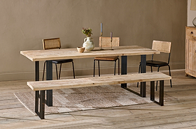 The Fia Dining Table by nkuku, crafted from sustainable mango wood with black metal legs, is styled with ceramics and flowers. Three wooden chairs and a matching bench surround the table on a textured rug in this neutral-toned space.