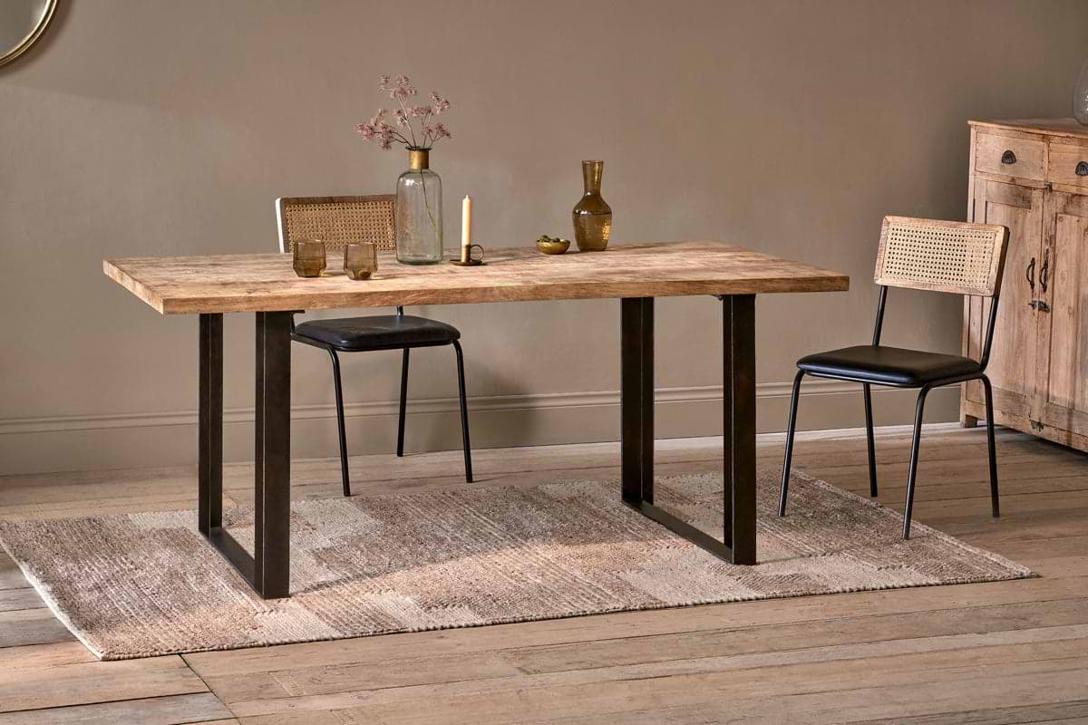 The Fia Dining Table by nkuku, crafted from sustainable mango wood with black metal legs, sits on a woven rug with cane-back chairs. Glass vases, dried flowers, and a small bowl adorn the table; a wooden cabinet stands against the beige wall.