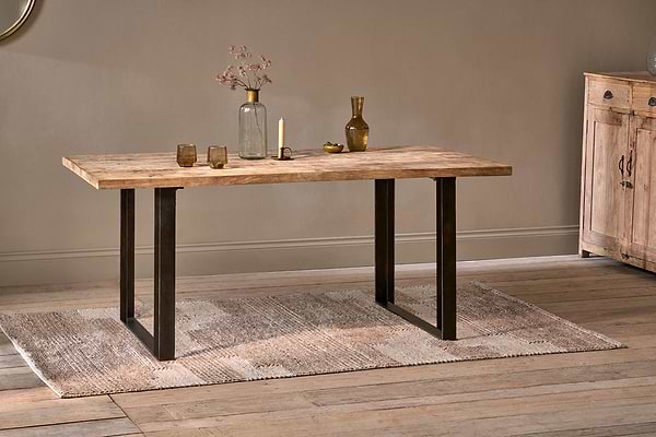 The nkuku Fia Dining Table, crafted from sustainable mango wood with black metal legs, stands on a textured beige rug and displays a glass vase of dried flowers, brass candle holder, gold bottle, and two low glasses on light wood floors.