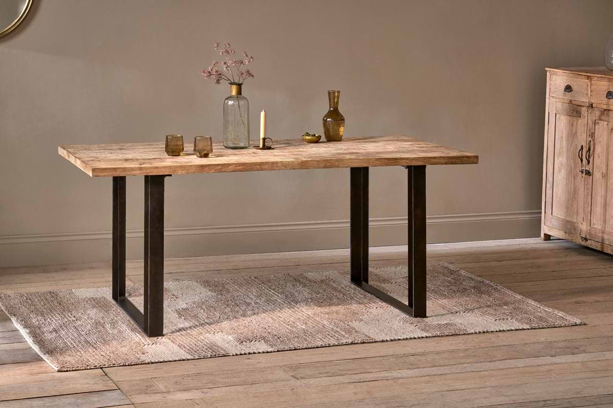 The nkuku Fia Dining Table, crafted from sustainable mango wood with black metal legs, stands on a textured beige rug and displays a glass vase of dried flowers, brass candle holder, gold bottle, and two low glasses on light wood floors.