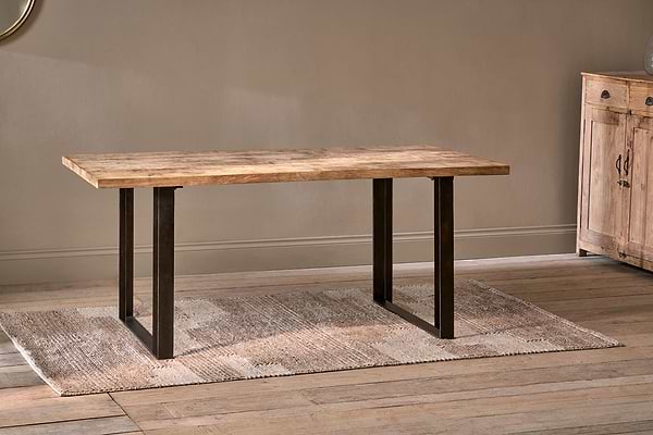 The Fia Dining Table by nkuku, featuring a rectangular mango wood top and black metal legs, stands on a beige woven rug in a room with light wood floors and neutral walls. A wooden cabinet is partially visible on the right.