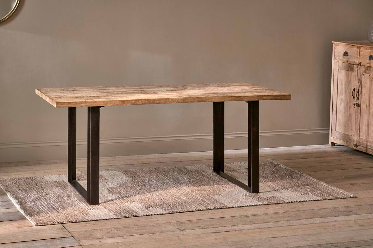 The Fia Dining Table by nkuku, featuring a rectangular mango wood top and black metal legs, stands on a beige woven rug in a room with light wood floors and neutral walls. A wooden cabinet is partially visible on the right.