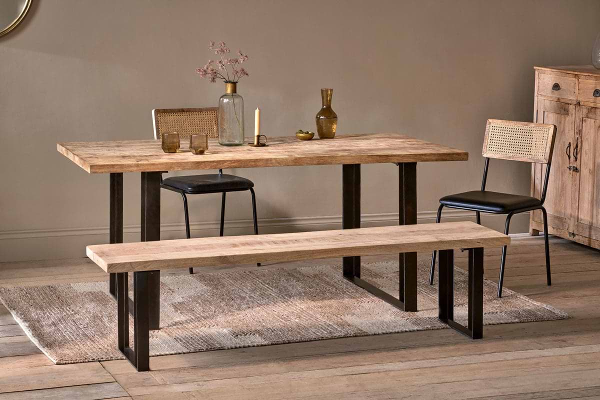 A dining space featuring the nkuku Fia Dining Table in sustainable mango wood with black metal legs, flanked by two cane-back chairs and a matching bench. The table is styled with glass vases, glasses, dried flowers; a sideboard and rug finish the look.