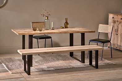 A dining space featuring the nkuku Fia Dining Table in sustainable mango wood with black metal legs, flanked by two cane-back chairs and a matching bench. The table is styled with glass vases, glasses, dried flowers; a sideboard and rug finish the look.