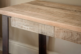 The nkuku Fia Mango Wood Desk - Natural features a slim mango wood tabletop with visible grain, paired with straight dark metal legs, shown against a beige wall and white baseboard.