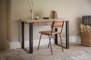 The nkuku Fia Mango Wood Desk - Natural features rustic charm, paired with a brown leather chair, a vase of dried flowers, books, and a basket of wheat stalks placed near a radiator.