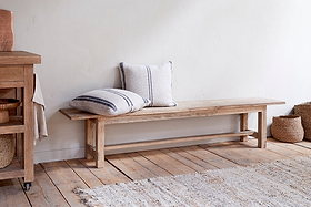 The nkuku Fioni Reclaimed Wood Bench - Natural sits against a white wall on wooden flooring, paired with two striped, neutral pillows. Woven baskets and a textured rug complete the cozy, minimalist farmhouse look.